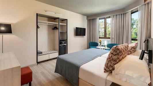 Hotellihuone, jossa on parivuode, kaksi sinistä tuolia pöydän ääressä ikkunoiden edessä sekä avohyllyillä varustettu vaatekaappi. (Tekoälyn luoma teksti)