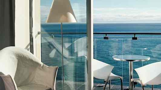 Näkymä huoneesta merelle, jossa on parveke lasikaiteilla, pöytä ja tuolit. (Tekoälyn luoma teksti)