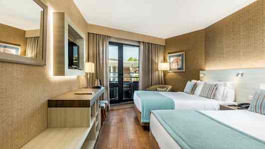 Hotellihuone, jossa on kaksi erillistä vuodetta, työpöytä, seinälle asennettu televisio ja ikkuna, joka johtaa parvekkeelle. (Tekoälyn luoma teksti)