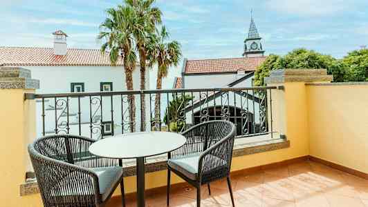 Kuvassa on parveke, jossa on kaksi tuolia ja pöytä. Näkymä valkoiseen taloon, jossa punaiset katot ja palmuja. (Tekoälyn luoma teksti)