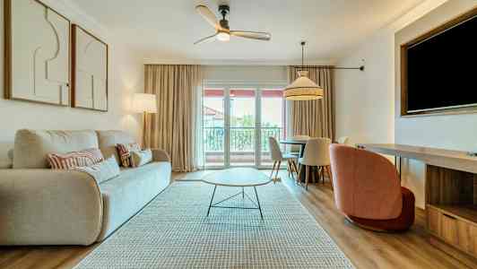 Olohuone, jossa on vaalea sohva, moderni sohvapöytä ja ruokapöytä tuoleineen. Ikkunasta verhojen kanssa pääsee päivänvaloa sisään. (Tekoälyn luoma teksti)