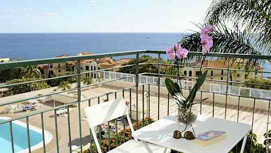 Näkymä parvekkeelta, jossa on pöytä ja tuolit sekä orkidea purkissa. Altaan ja rakennusten taustalla näkyy meri. (Tekoälyn luoma teksti)