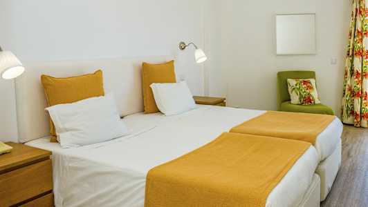 Hotellihuone, jossa kaksi erillistä sänkyä ja keltaiset vuodevaatteet. Vihreä nojatuoli kukikkaalla tyynyllä ja verhot taustalla. (Tekoälyn luoma teksti)