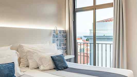 Hotellihuone, jossa on parisänky, valkoiset vuodevaatteet ja siniset tyynyt, ikkunan vieressä näkymän kera. (Tekoälyn luoma teksti)