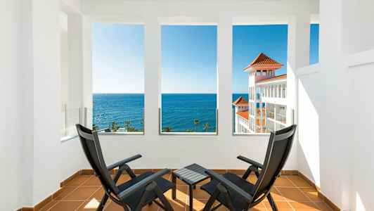 Kaksi mustaa tuolia ja pöytä terassilla, josta on näkymä merelle ja valkoiseen rakennukseen, jossa on punaiset kattot. (Tekoälyn luoma teksti)