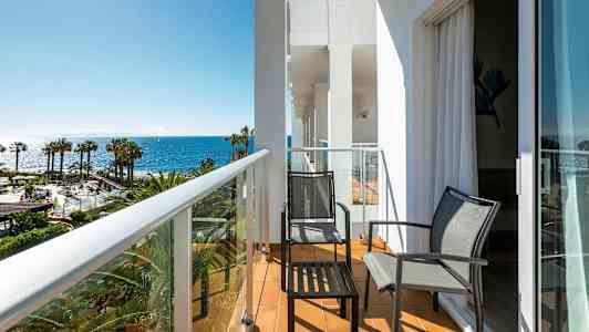 Parveke, jossa kaksi tuolia ja pöytä, näkymä palmuihin ja merelle. (Tekoälyn luoma teksti)