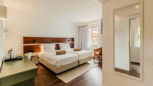 Hotellihuone, jossa on kaksi erillistä vuodetta, työpöytä ja seinäpeili. Huoneessa on suuret ikkunat verhoilla. (Tekoälyn luoma teksti)