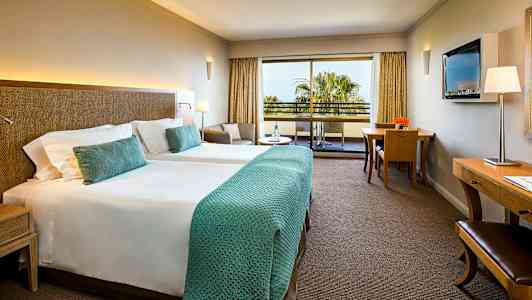 Hotellihuone, jossa on kaksi erillistä vuodetta, istuinryhmä, työpöytä ja parveke näkymällä. (Tekoälyn luoma teksti)