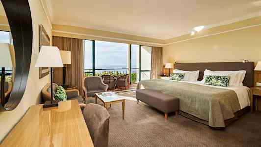 Hotellihuone, jossa on sänky, istuinryhmä ja näkymä parvekkeelle ja merelle. (Tekoälyn luoma teksti)