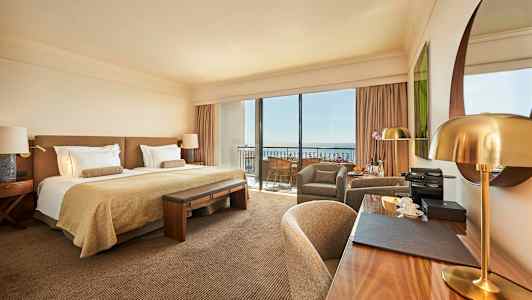 Tyylikäs hotellihuone, jossa on parivuode, kirjoituspöytä ja istuinryhmä, sekä suuret ikkunat parvekkeelle. (Tekoälyn luoma teksti)