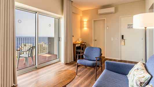 Moderni hotellihuone, jossa istuinpaikkoja ja näkymä merelle suurten ikkunoiden läpi. (Tekoälyn luoma teksti)