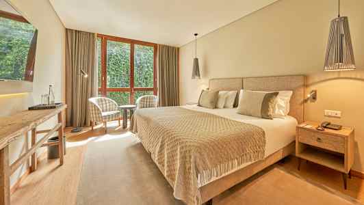 Hotellihuone, jossa on leveä sänky, kaksi nojatuolia ikkunan ääressä ja pöytä. (Tekoälyn luoma teksti)