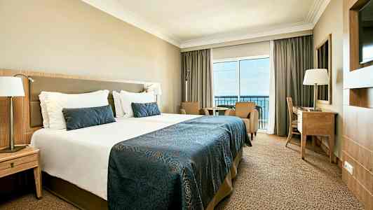 Hotellihuoneen kuva, jossa on suuri sänky, kaksi nojatuolia, kirjoituspöytä ja parveke merinäköalalla. (Tekoälyn luoma teksti)