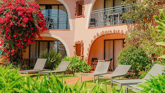 Rakennus, jossa on kaarevat parvekkeet vihreyden ja kukkien keskellä, aurinkotuoleja nurmikolla. (Tekoälyn luoma teksti)