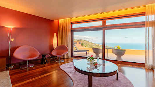 Huone, jossa punaiset seinät, kaksi tuolia, pöytä ja suuret ikkunat, jotka avautuvat terassille merinäköalalla. (Tekoälyn luoma teksti)