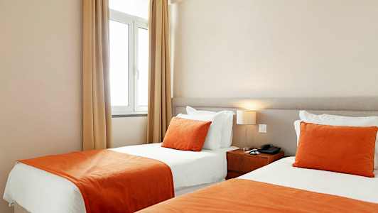 Hotellihuone, jossa on kaksi vierekkäistä yhden hengen vuodetta, valkoiset lakanat ja oranssit päiväpeitteet sekä koristetyynyt. (Tekoälyn luoma teksti)