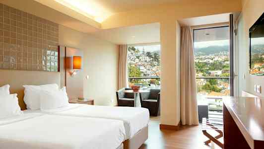 Hotellihuone, jossa kaksi erillistä vuodetta, oleskelutila ikkunan ääressä, josta näkymät vuoristoiselle alueelle kaupunkiin. (Tekoälyn luoma teksti)