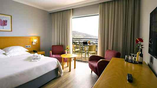 Hotellihuone, jossa on parivuode, kaksi punaista nojatuolia, kirjoituspöytä juomien ja lasien kanssa sekä parveke maisemilla. (Tekoälyn luoma teksti)