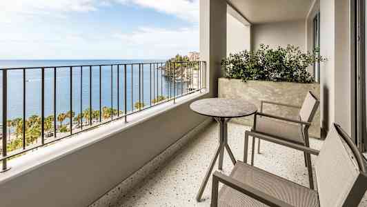 Parveke, jossa on kaksi tuolia ja pöytä sekä näkymä merelle ja palmuihin. (Tekoälyn luoma teksti)