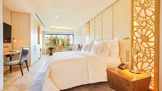 Moderni hotellihuone, jossa suuri sänky, työpöytä, tuoli ja parveke, jolta on näkymä. (Tekoälyn luoma teksti)