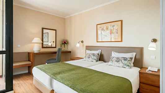 Kuva hotellihuoneesta, jossa on kaksi yksittäistä vuodetta, valkoiset vuodevaatteet ja vihreät päiväpeitot sekä taustalla työpöytä ja tuoli peilin kera. (Tekoälyn luoma teksti)