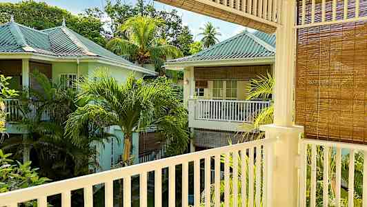 Näköala parvekkeelta, josta näkyy vehreä alue pastellinvärisine taloineen ja palmupuineen. (Tekoälyn luoma teksti)