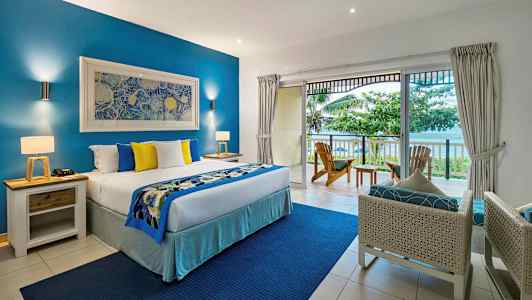 Hotellihuone, jossa on suuri sänky, sinisiä ja keltaisia koristeellisia tyynyjä, parveke merinäköalalla ja ympäröivää vehreyttä. (Tekoälyn luoma teksti)