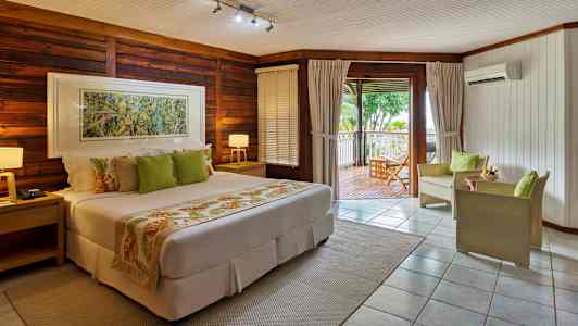 Hotellihuone lämpimin sävyin, jossa on sänky, istuimet ja terassi, jolta avautuu näkymä vehreään maisemaan. (Tekoälyn luoma teksti)