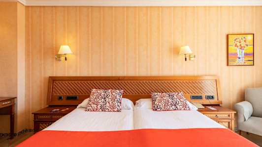 Hotellihuone, jossa on kahden hengen sänky, valkoiset ja oranssiset vuodevaatteet sekä koristeelliset tyynyt. Sängyn pääty on tehty puusta ja siinä on ruutukuviointia. (Tekoälyn luoma teksti)