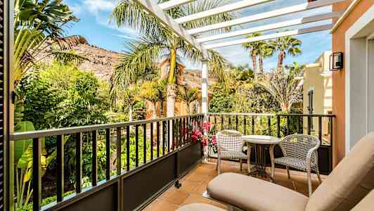 Parveke, jossa kaksi aurinkotuolia, pöytä ja tuolit. Näkymä vehreään puutarhan ja vuoriin. (Tekoälyn luoma teksti)