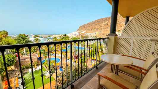 Näkymä parvekkeelta uima-allasalueelle, jossa on vehreyttä, palmuja ja taustalla näkyvä vuoren rinne. (Tekoälyn luoma teksti)