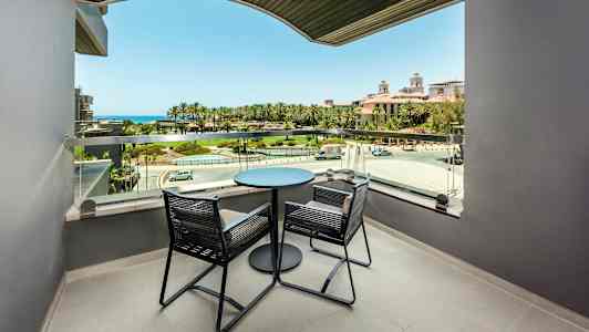 Parveke, jossa on kaksi tuolia ja pöytä, näkymä vihreälle alueelle ja merelle. (Tekoälyn luoma teksti)