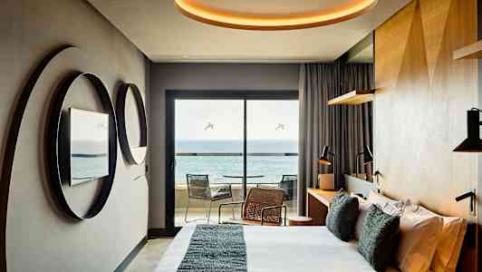 Moderni hotellihuone, jossa on sänky, peilit seinällä, kirjoituspöytä ja parveke merinäköalalla. (Tekoälyn luoma teksti)