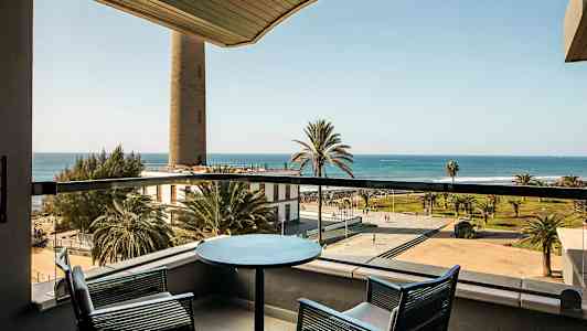 Näkymä parvekkeelta, jossa on kaksi tuolia ja pöytä, kohti rantapromenadia, palmuja ja merta. (Tekoälyn luoma teksti)