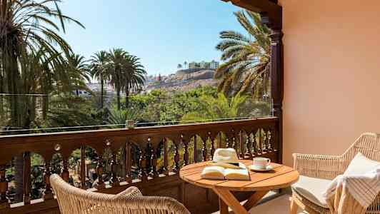 Terassi, jossa pöytä, kirja ja aurinkohattu, näkymä palmuihin ja rakennuksiin. (Tekoälyn luoma teksti)