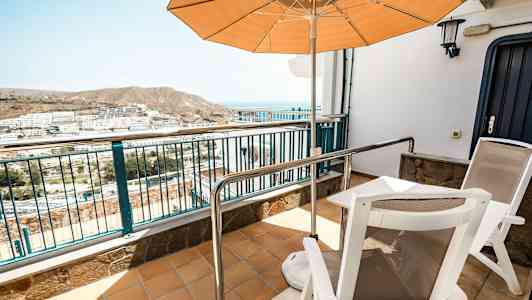 Terassi, jossa on kaksi tuolia, pöytä ja aurinkovarjo, näkymä asuinalueelle ja vuoristoon taustalla. (Tekoälyn luoma teksti)