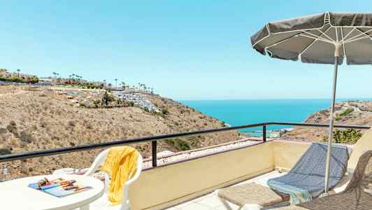 Näköalapaikka, jossa on pöytä ja tuoleja, aurinkovarjo sekä aurinkotuoleja katsellen rannikkoaluetta ja merta. (Tekoälyn luoma teksti)