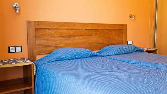 Sänky sinisillä pilkullisilla vuodevaatteilla ja puinen sängynpääty oranssia seinää vasten. (Tekoälyn luoma teksti)