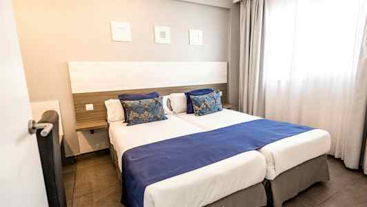 Hotellihuone, jossa on kaksi vierekkäistä yhden hengen vuodetta, siniset päiväpeitteet ja koristeelliset tyynyt. (Tekoälyn luoma teksti)