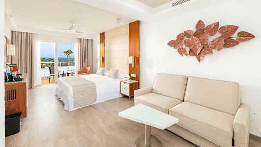 Valoisa hotellihuone, jossa on sänky, sohva ja parveke sekä merinäköala. (Tekoälyn luoma teksti)