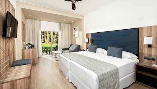 Moderni hotellihuone, jossa on suuri sänky, istuinalueita ja ikkunat näkymällä vehreään puutarhaan. (Tekoälyn luoma teksti)