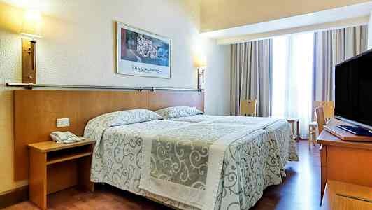 Hotellihuone, jossa on parivuode kuviollisilla päiväpeitteillä, seinävalaisimia ja televisio kirjoituspöydällä. Istuimia ja verhoilla varustettu ikkuna ovat myös mukana. (Tekoälyn luoma teksti)