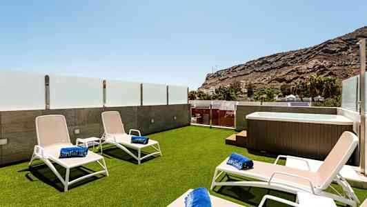 Terassi, jossa aurinkotuoleja, kylpypyyhkeitä ja poreallas, lasikaiteiden ympäröimä ja näkymä vuorille. (Tekoälyn luoma teksti)