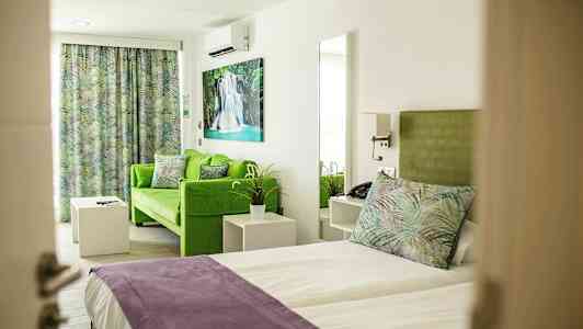 Huone, jossa sänky ja vihreä sohva, värikkäitä tyynyjä ja verhoja sekä taidetta seinällä. (Tekoälyn luoma teksti)