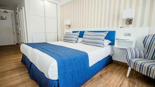 Tyylikäs hotellihuone, jossa on parivuode sinisillä ja valkoisilla vuodevaatteilla sekä raidalliset tyynyt, vieressä raidallinen nojatuoli. (Tekoälyn luoma teksti)