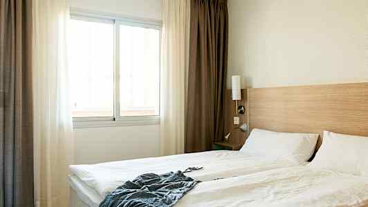 Hotellihuone, jossa on parivuode, valkoiset vuodevaatteet, puinen sängynpääty ja ikkuna vaaleilla verhoilla. (Tekoälyn luoma teksti)