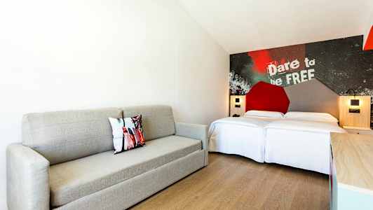 Moderni hotellihuone, jossa harmaa sohva, parivuode ja koristeellinen seinä tekstillä. (Tekoälyn luoma teksti)