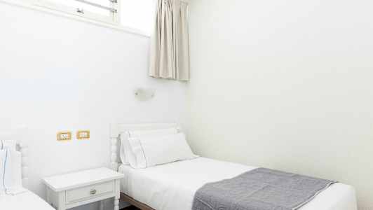 Kaksi erillistä sänkyä, joissa on valkoiset vuodevaatteet ja harmaa peite, sijaitsevat valoisassa huoneessa. (Tekoälyn luoma teksti)