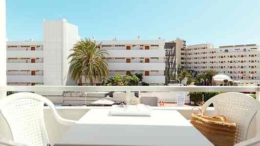 Näkymä parvekkeelta, jossa istuinryhmä, rakennuksia ja palmuja näkyvissä taustalla kirkkaan sinisen taivaan alla. (Tekoälyn luoma teksti)