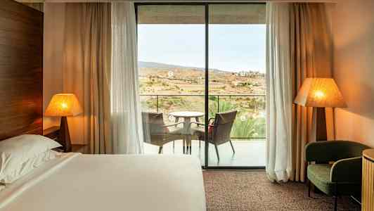 Hotellihuone, jossa on vuode, kaksi pöytälamppua, nojatuoli ja parveke istuimineen. (Tekoälyn luoma teksti)